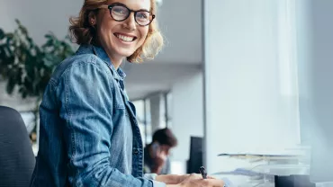 Personne souriante portant des lunettes, assise à un bureau moderne.