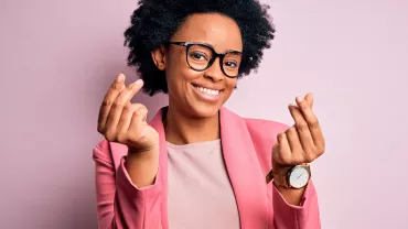 Personne souriante portant des lunettes et un blazer rose, posant devant un arrière-plan rose clair.