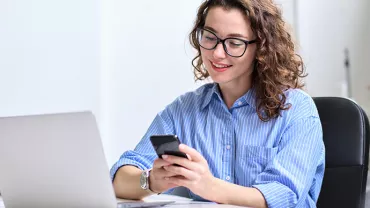 Personne souriante utilisant un smartphone devant un ordinateur portable.