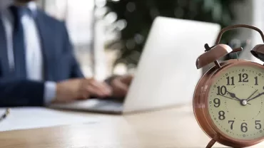 Réveil ancien sur un bureau avec une personne utilisant un ordinateur portable en arrière-plan.