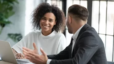 Deux personnes souriant et discutant à un bureau avec un ordinateur portable.