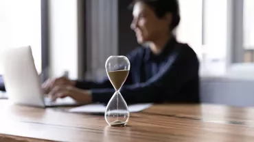 Une personne travaille sur un ordinateur portable. Un sablier repose sur le bureau au premier plan.
