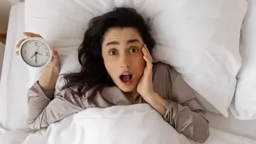 Femme dans son lit tenant son réveil et réalisant qu'elle est en retard