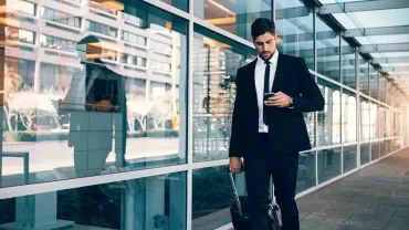 Homme regardant son téléphone tout en tirant une valise à roulette dans la rue.
