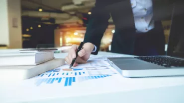 Personne écrivant sur des documents avec un graphique à côté d'un ordinateur portable.