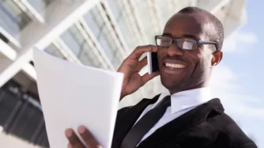 Personne souriant au téléphone tout en tenant des documents à l'extérieur d'un bâtiment moderne.