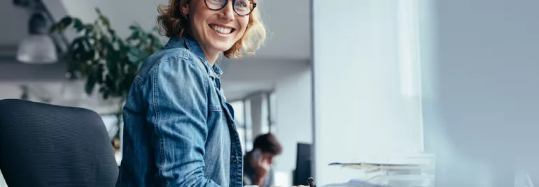 Personne souriante portant des lunettes, assise à un bureau moderne.