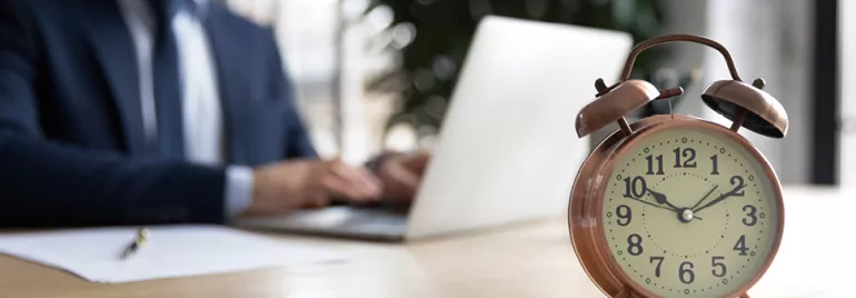 Réveil ancien sur un bureau avec une personne utilisant un ordinateur portable en arrière-plan.