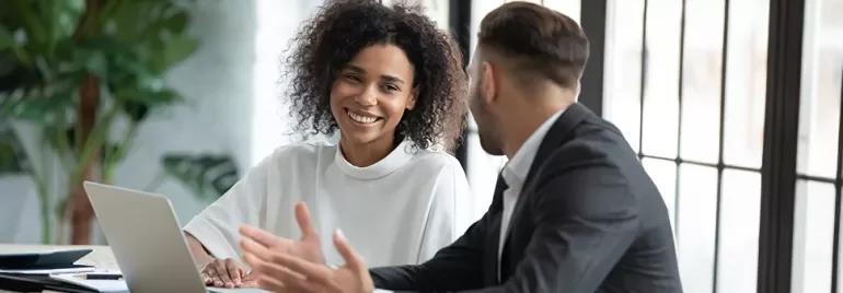 Deux personnes souriant et discutant à un bureau avec un ordinateur portable.