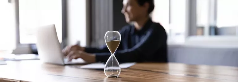 Une personne travaille sur un ordinateur portable. Un sablier repose sur le bureau au premier plan.