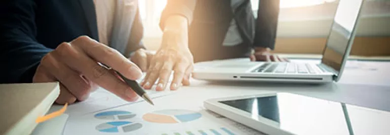 Deux personnes discutant de graphiques financiers sur un bureau avec un ordinateur portable et un smartphone.
