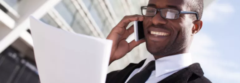 Personne souriant au téléphone tout en tenant des documents à l'extérieur d'un bâtiment moderne.
