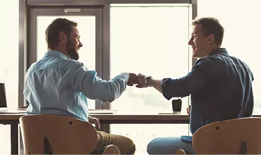 Deux personnes se saluent avec un geste de poing, assises à une table près d'une fenêtre.