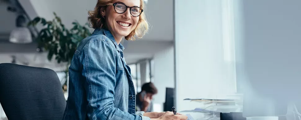Personne souriante portant des lunettes, assise à un bureau moderne.