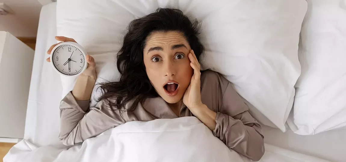 Femme dans son lit tenant son réveil et réalisant qu'elle est en retard