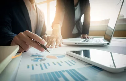 Deux personnes discutant de graphiques financiers sur un bureau avec un ordinateur portable et un smartphone.